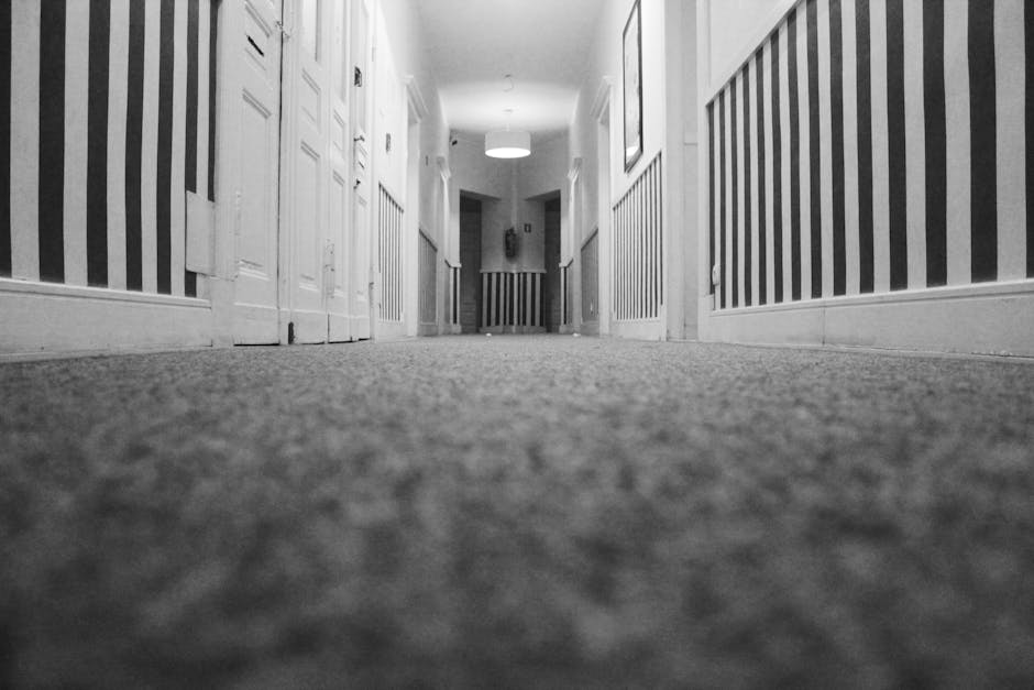 A professional cleaner dressed in full white protective gear, including a disposable suit, gloves, and shoe covers, is using a vacuum or extraction machine to deep clean a beige carpet in a bright, mo