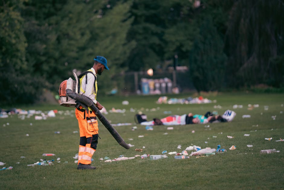 A sanitation worker wearing a blue cap, yellow high-visibility jacket, and orange trousers with reflective strips is using a leaf blower to clear litter from a grassy park area, likely during or after